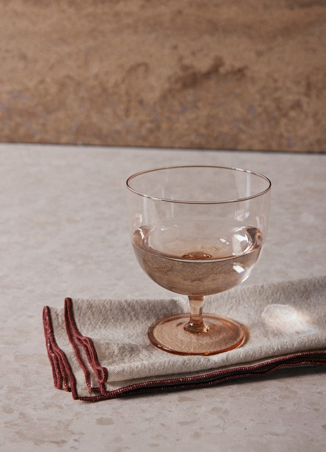 A clear glass with a short stem, filled with water, sits on a folded beige cloth with red trim—an elegant example of chic homeware under £50—placed on a light stone surface against a textured brown background.