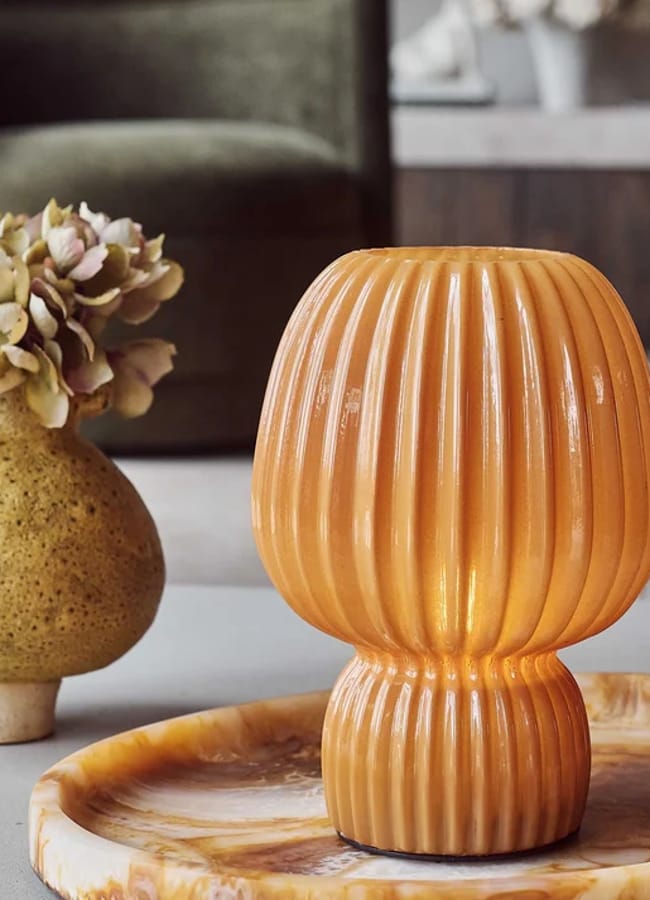 A ribbed orange glass lamp glows on a marble tray next to a round yellow vase holding dried hydrangea flowers, with caramel homeware accents and a blurred green chair and decor in the background.