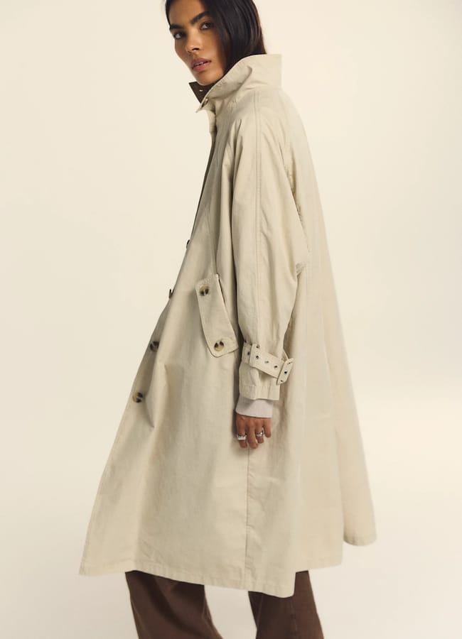 A person showcasing corporate looks for women is wearing a light beige trench coat with the collar up, standing sideways and looking at the camera. The coat has button details, paired effortlessly with brown trousers. The background is plain and light-coloured.