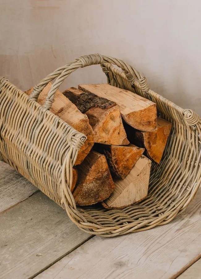 A wicker basket filled with chopped firewood sits on a wooden floor next to a light-coloured wall, offering a touch of chic homeware under £50 to elevate your living space.