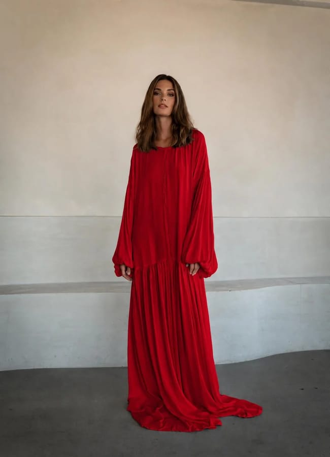 A woman stands indoors wearing a long, loose-fitting red dress with long sleeves—a timeless piece often seen in collections from small fashion brands. The background is plain and neutral, with soft lighting.