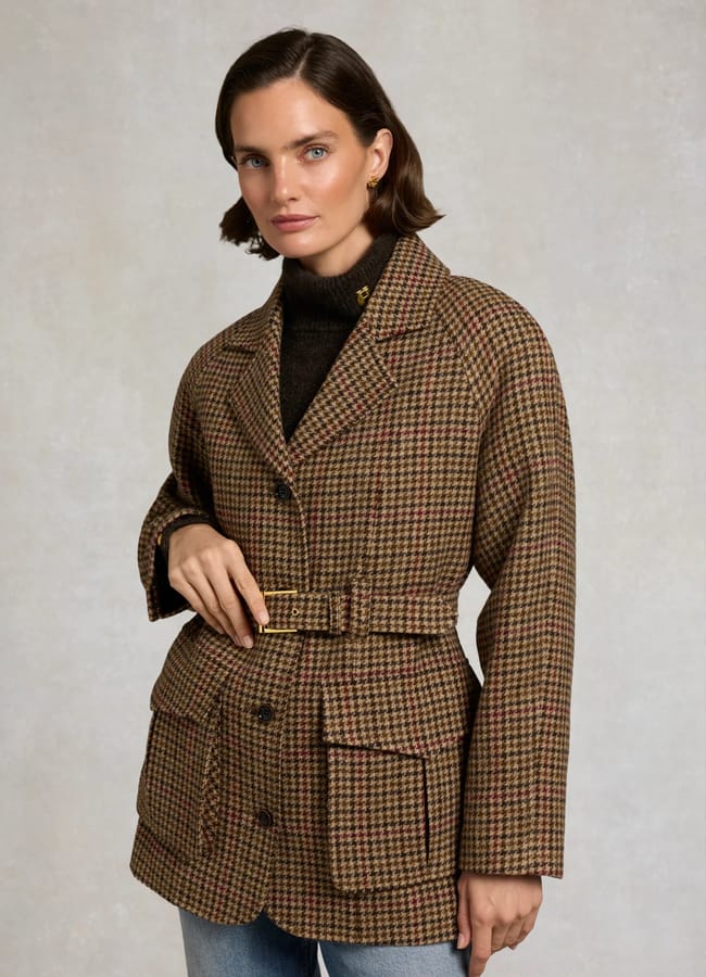 Woman wearing a brown checked belted blazer, styled as one of this season’s must-have lightweight jackets, over a dark polo neck and light blue jeans, standing against a plain light background.