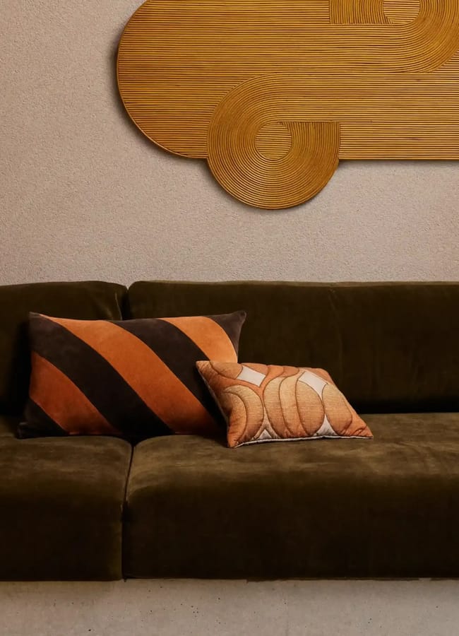A dark green velvet sofa with two decorative cushions, one striped in brown and orange, the other with a curved pattern, creates an inviting focal point. Above it, a textured, abstract wooden wall art piece hangs against a beige wall—an elegant reminder that chic homeware under £50 can transform any space with style.