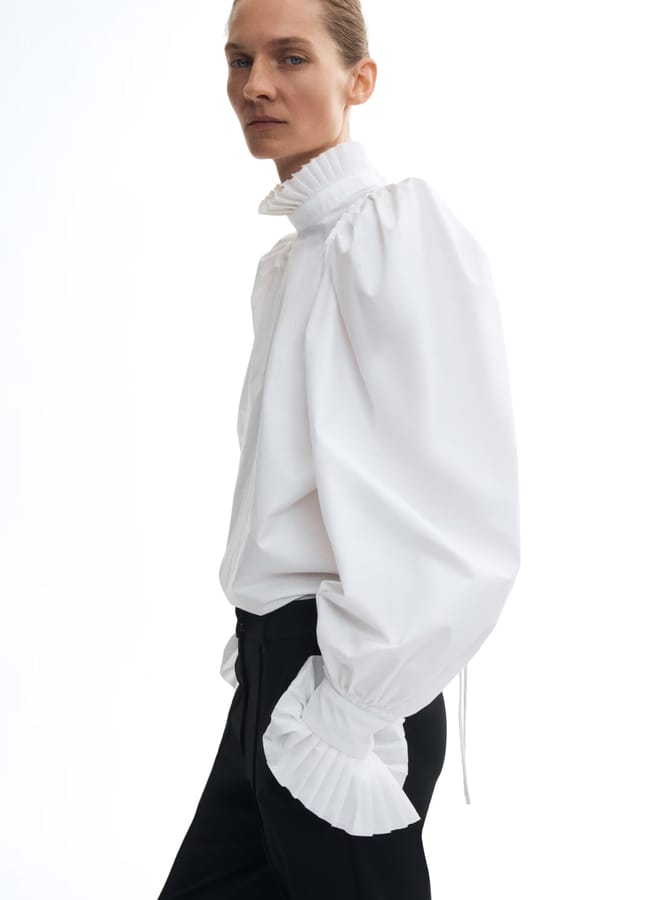 A person wearing a white blouse with voluminous pleated sleeves and cuffs, paired with black trousers, showcases effortless fashion against a plain white background.