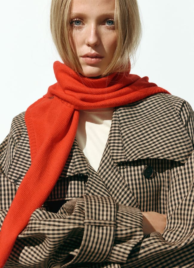 A person with blonde hair wears a brown checked coat, a cream shirt, and a bright red scarf draped around their neck—timeless fashion essentials perfect for an effortless wardrobe upgrade—standing against a plain light background.
