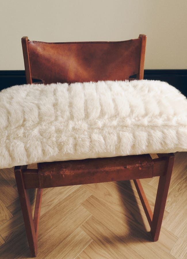 A brown leather chair with a wooden frame has a fluffy white cushion draped over the seat. Perfect for showcasing chic homewares, the chair is placed on a wooden floor against a cream-coloured wall.