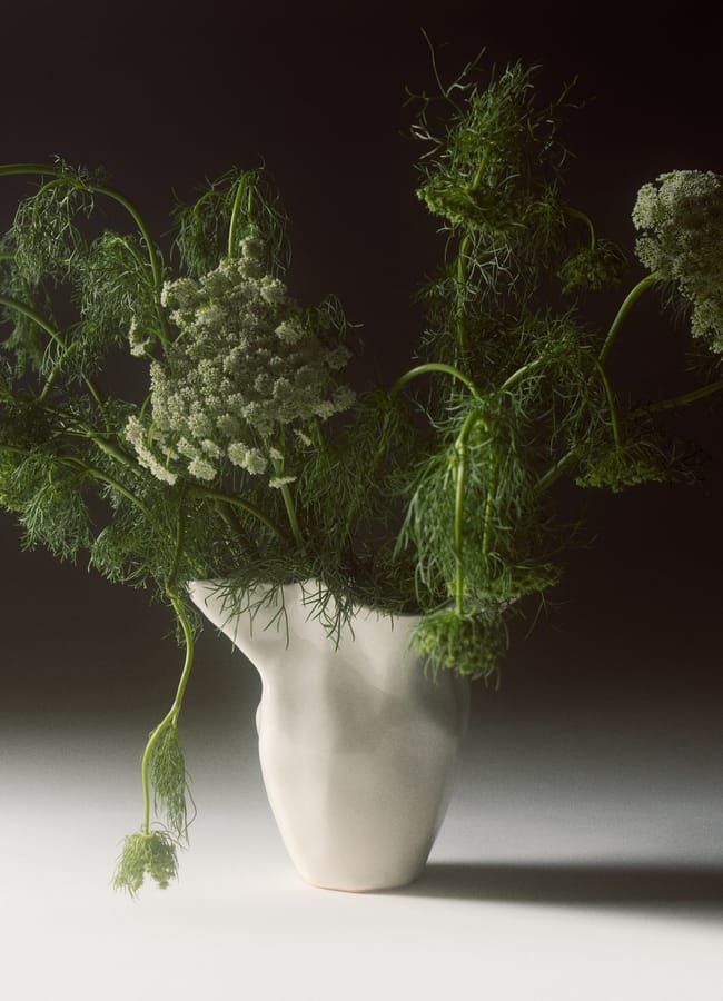 A chic homeware under £50, this white, abstract-shaped vase holds green, feathery foliage and delicate white flowers, set against a dark background with soft lighting.