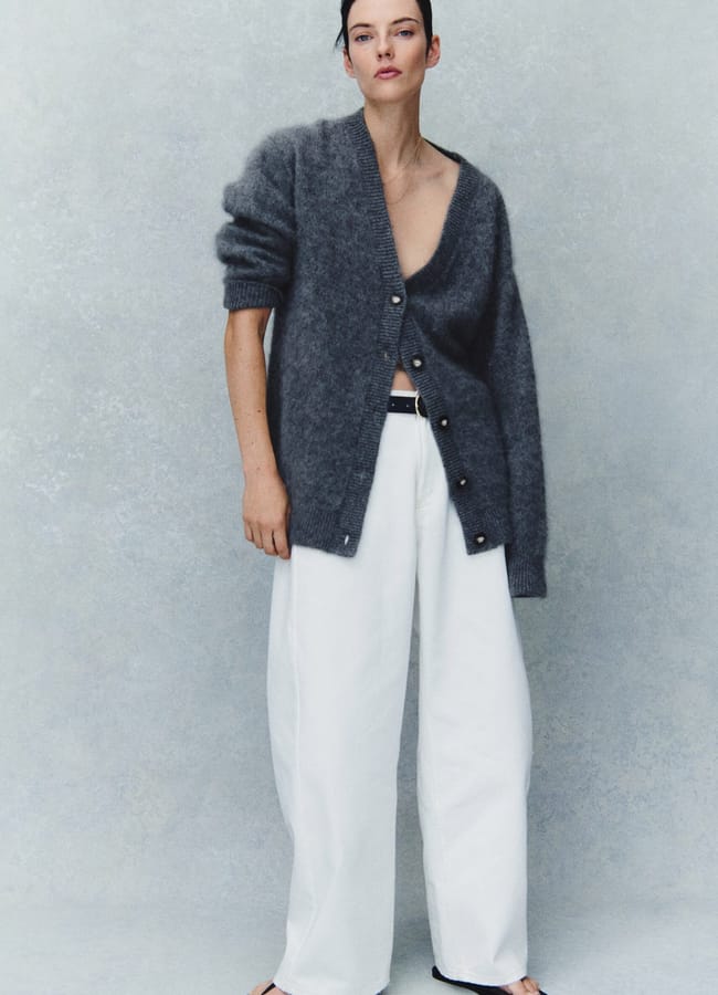 A person wearing a loose grey cardigan with large buttons, white wide-leg trousers, and black ballet pumps stands against a light grey textured background—a look that perfectly captures the clean lines and effortless sophistication often found in minimalist high street brands.