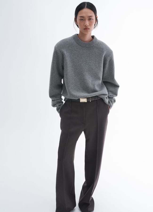 Person wearing a grey jumper and dark wide-leg trousers, styled in the spirit of minimalist high street brands, stands with hands in pockets against a plain white background, looking directly at the camera with a neutral expression.