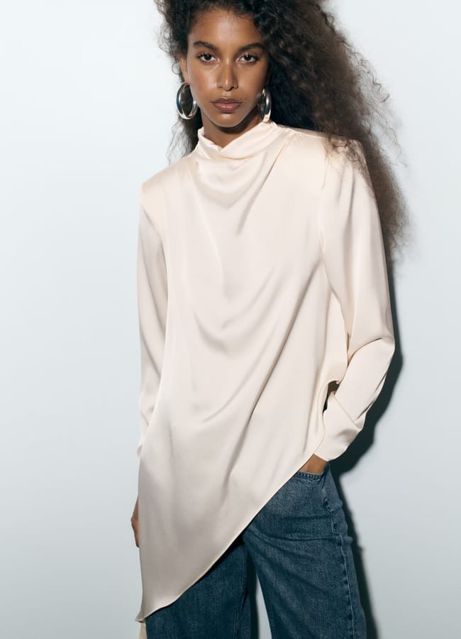 A woman with long curly hair wearing a cream silk asymmetrical blouse and blue jeans from minimalist high street brands stands against a plain light background, looking at the camera.