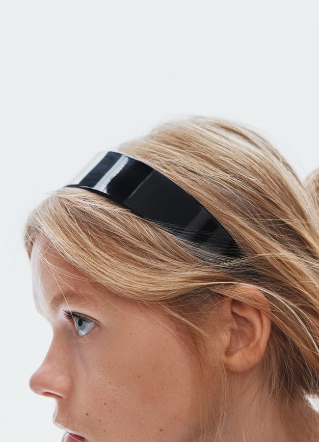 A close-up side profile of a young person with light skin and blonde hair wearing a wide, shiny black hairband, set against a plain white background—a look reminiscent of minimalist high street brands.