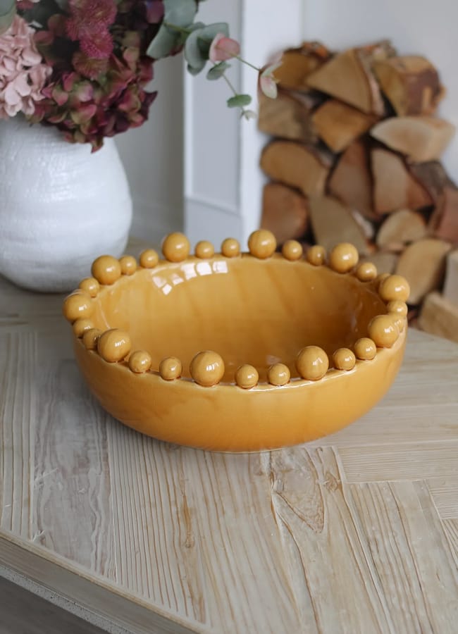 A yellow ceramic bowl with round decorative beads around the rim sits on a light wooden table, adding a touch of caramel homeware charm to the scene. In the background are stacked firewood and a white vase with pink and green flowers.