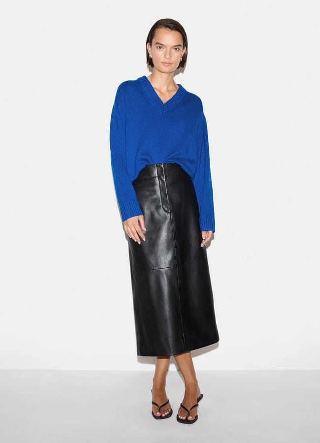 A woman stands against a white background wearing a vibrant International Klein Blue jumper, a black leather midi skirt, and black heeled sandals. She has short brown hair and a relaxed, confident posture, embodying bold colour everywhere in her look.