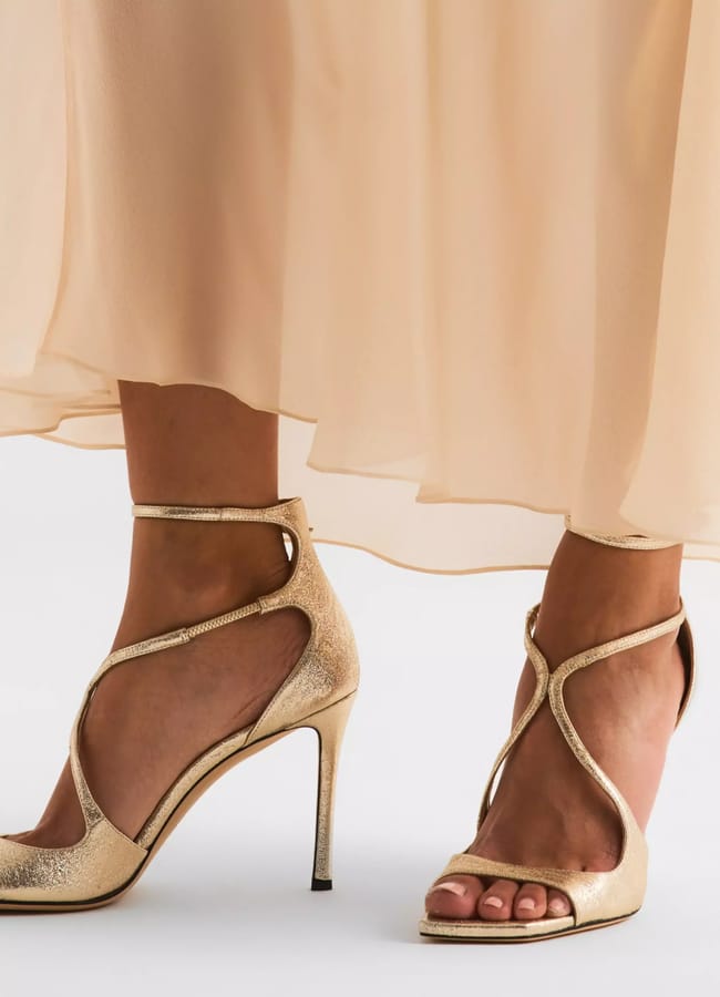 A person wearing sexy heels—gold strappy high-heeled sandals with an ankle strap—paired with a flowing, light beige dress that falls to mid-calf.