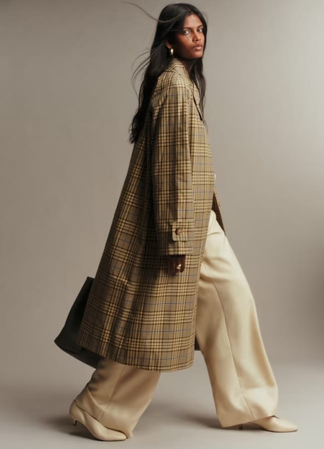 A woman in a long checked coat, wide-leg cream trousers, and cream heels poses against a neutral background, carrying a large black bag—a striking example of new season fashion.