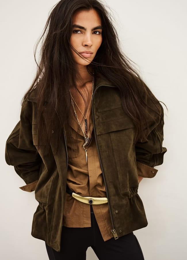 A woman with long dark hair wears a lightweight olive green jacket over a brown shirt, black trousers, layered necklaces, and a gold belt, standing against a plain white background.