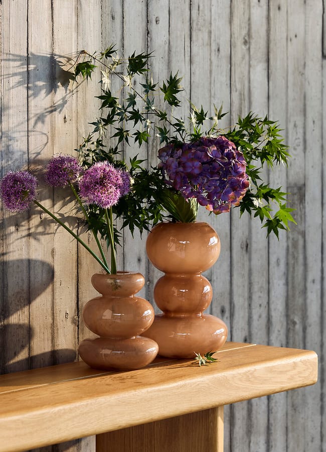 Two glossy, stacked ceramic vases with pinkish tones hold purple flowers and green leaves, sitting on a wooden bench against a light-coloured wooden plank wall. Shadows fall across the scene, beautifully highlighting this chic homeware under £50.