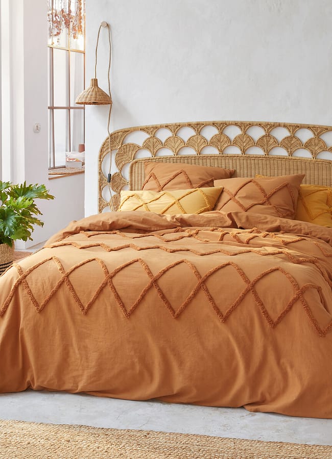 A cosy bedroom with a rattan headboard, mustard-coloured bedding with a diamond pattern, matching pillows, hanging woven lamps, touches of caramel homeware, a large green potted plant, and sunlight streaming through a window.