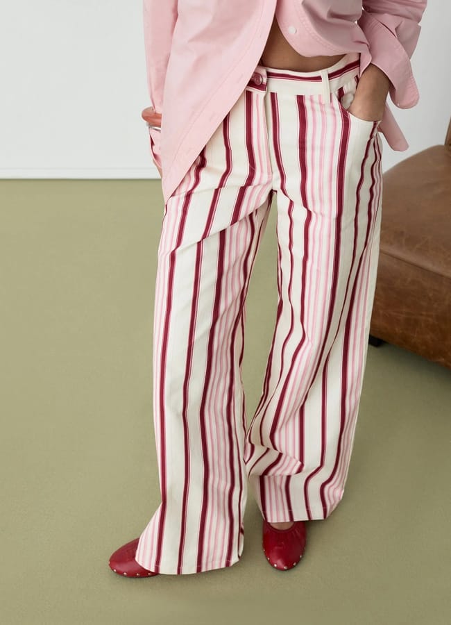 A person showcasing stylish fashion stands with hands in pockets on a light green floor, wearing a light pink shirt, wide-leg white trousers with pink and red vertical stripes, and red pointed shoes—proving that boyfriend-inspired clothes can be both chic and bold.