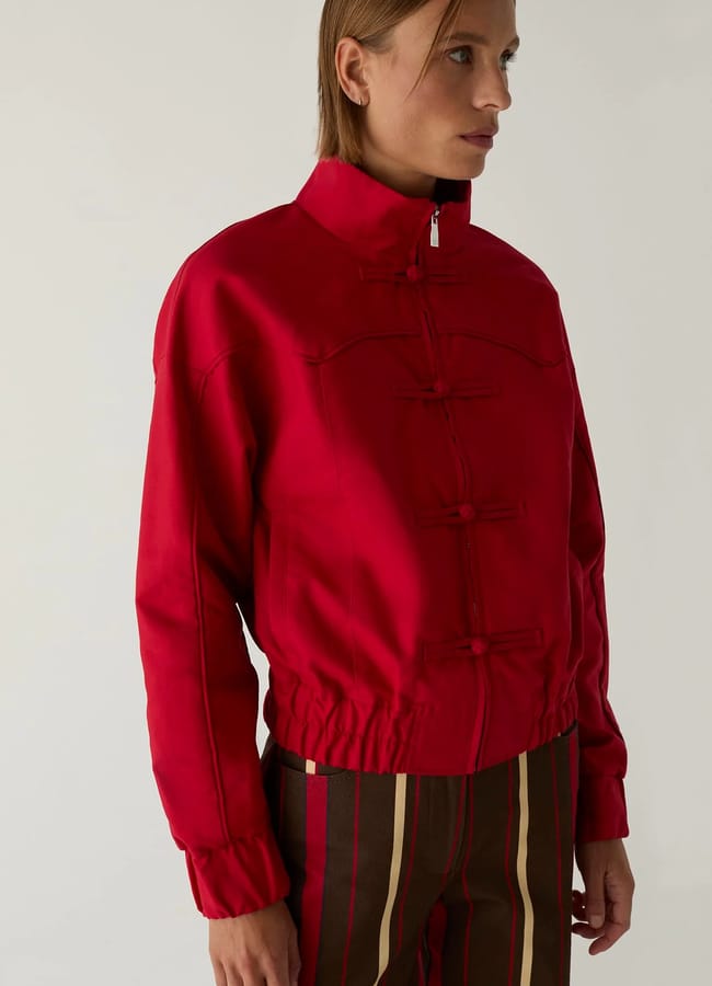 Person wearing a bright red, high-collared lightweight jacket with toggle fastenings and striped brown trousers, standing against a plain light background.