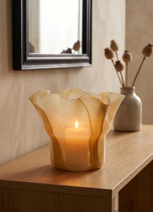 A lit candle inside a wavy, translucent glass holder sits on a wooden table. In the background, there is a beige vase with dried flowers and a framed mirror on the wall.