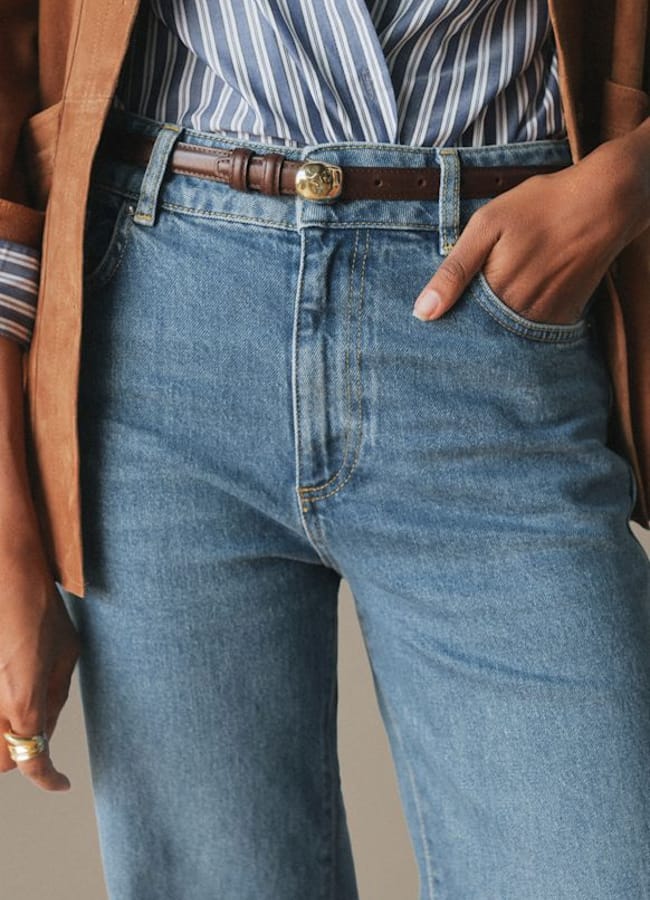 A person wearing blue jeans, a brown belt, a striped shirt, and a brown jacket stands with one hand in their pocket—perfectly embodying fashion essentials for an expensive-looking wardrobe upgrade.