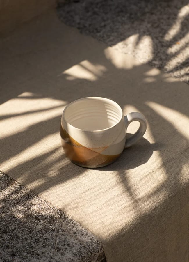 A caramel homeware touch is evident in the ceramic mug with earth-toned geometric patterns that sits on a beige cloth, surrounded by delicate shadows of leaves cast by sunlight.