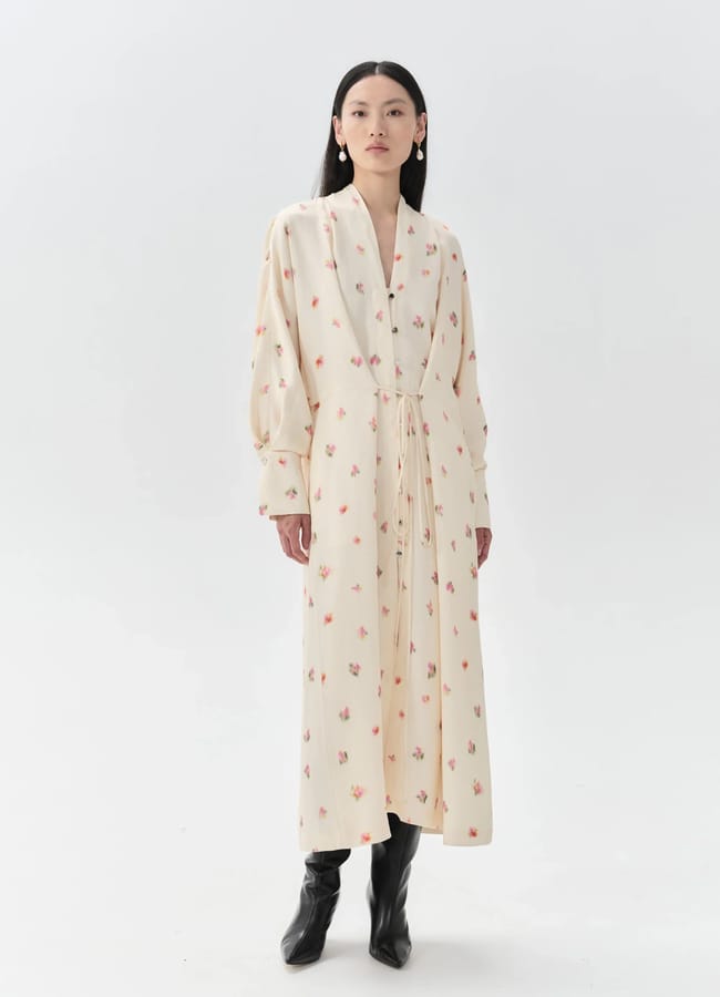A woman stands against a white background wearing a long, cream-coloured dress with a subtle floral pattern from one of her favourite small fashion brands. The dress features long sleeves, and she completes the look with black boots.