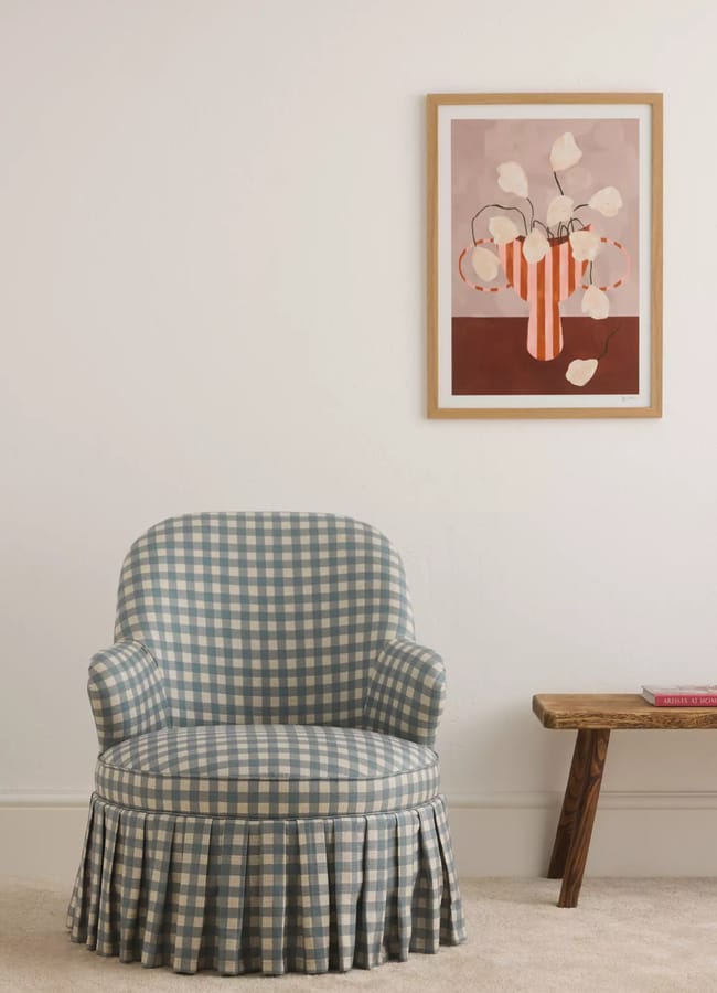 A blue and white chequered armchair with a skirt sits next to a wooden bench, beneath a framed abstract flower painting on a white wall, showcasing unexpected colour combinations that align with current colour trends.