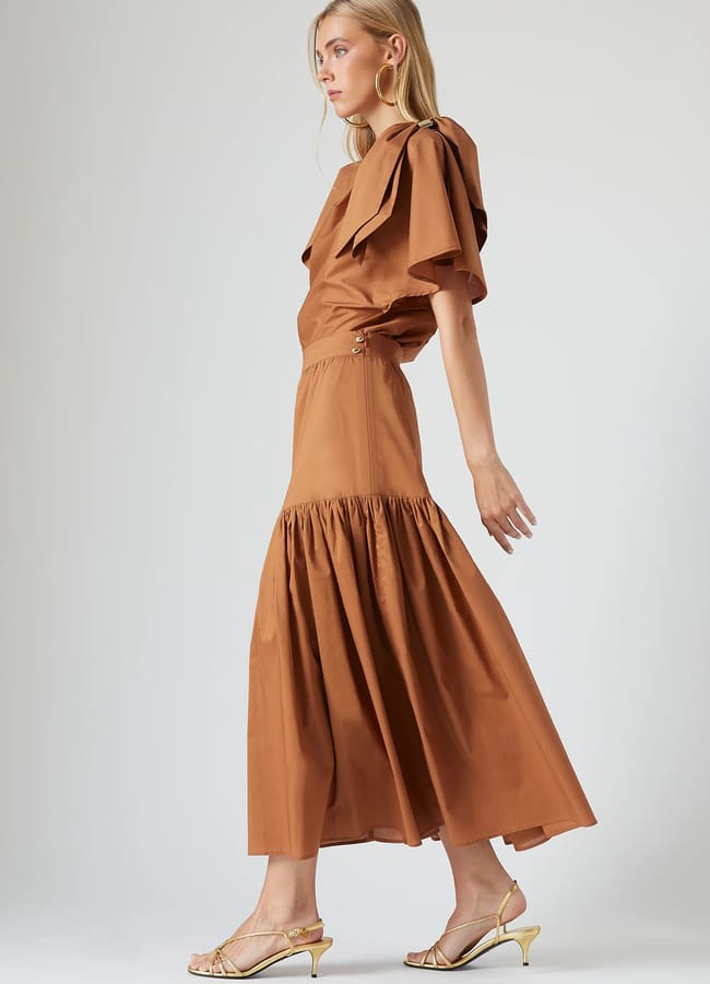 A woman in a brown, ruffled, short-sleeve midi dress from one of today’s small fashion brands and gold heels stands in profile against a plain background.