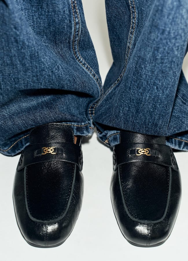 A close-up of a person wearing blue jeans and black leather loafers with gold hardware on the front, standing on a white surface—an effortless example of wardrobe essentials for those seeking affordable fashion.