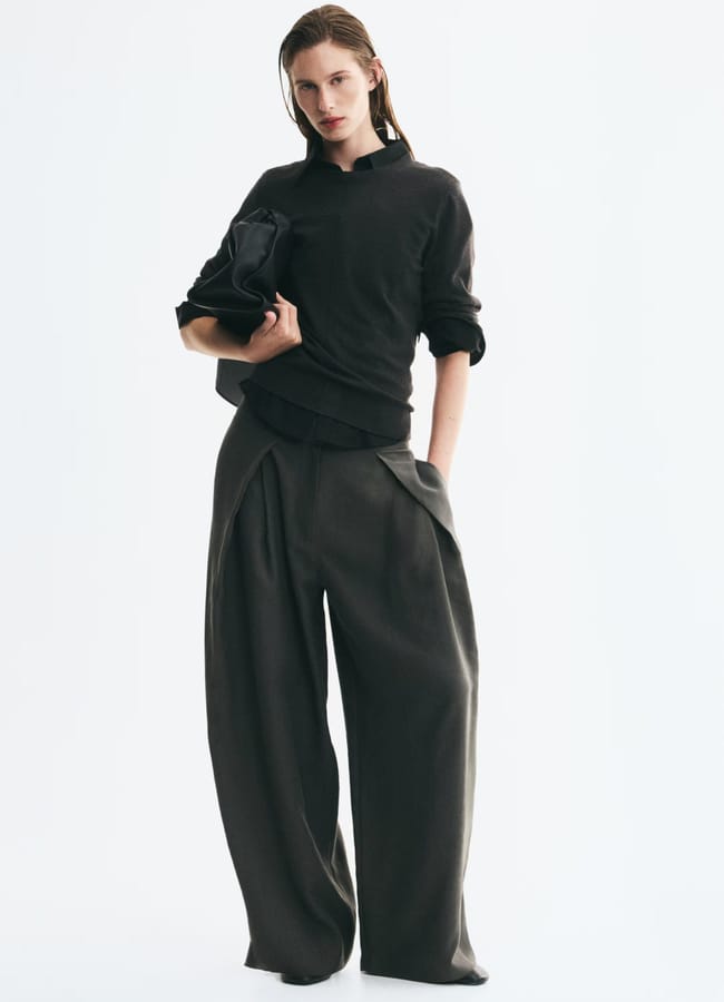 Person standing against a white background, wearing a dark, long-sleeve top with sleeves rolled up and wide-leg dark trousers—a look that embodies the effortless style found in minimalist high street brands. They hold a matching jacket and have a relaxed, confident posture with one hand in their pocket.