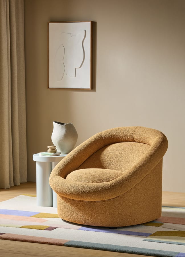 A modern, rounded beige armchair sits on a patterned rug beside two nesting tables holding a ceramic vase and subtle caramel homeware accents. Neutral curtains and abstract wall art create a cosy, minimalist living space.