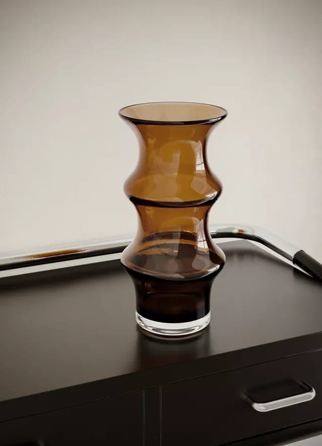 A brown, hourglass-shaped glass vase—a perfect piece of home décor—sits on a dark cupboard with two drawers, beside a metal bar with a black handle, against a plain light-coloured wall.