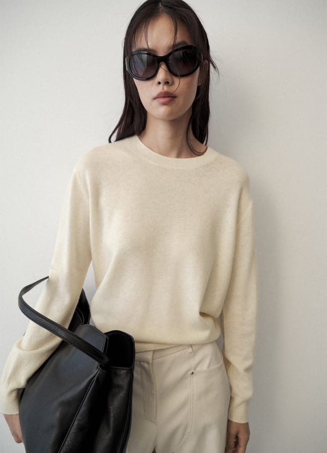 A person wearing black sunglasses, a cream jumper, and light trousers from minimalist high street brands stands against a plain white wall while carrying a large black bag over their shoulder.