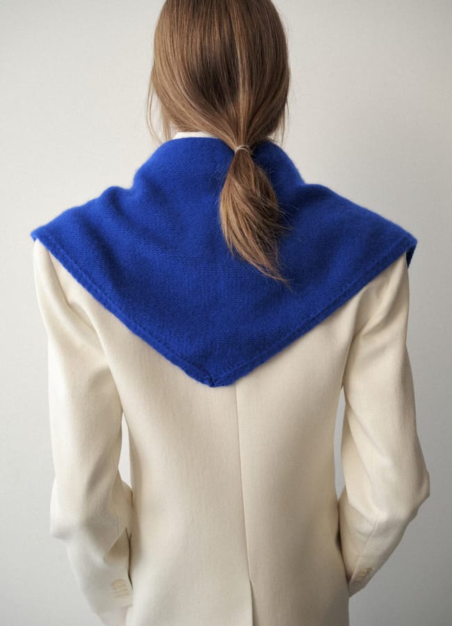 Person with light brown hair in a low ponytail, wearing a cream-coloured blazer and a large shawl in International Klein Blue draped over their shoulders, seen from behind against a plain background—a striking blend of colour everywhere in this minimalist scene.