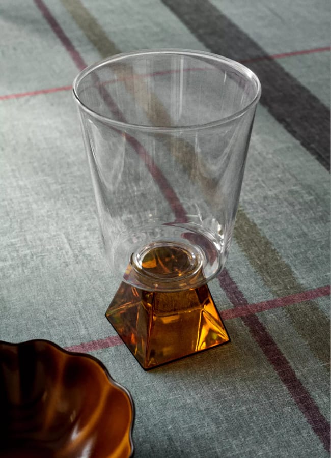 A clear glass with a geometric amber-coloured base sits on a textured grey tablecloth beside the edge of a brown, scalloped dish—an elegant example of chic homeware under £50.