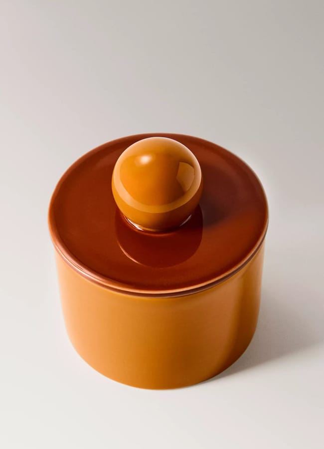 A glossy, cylindrical orange ceramic container from the Caramel Homeware collection, complete with a matching lid topped by a round knob, set against a plain light background.