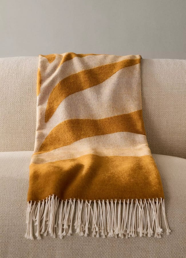 A cream and mustard yellow throw blanket with wavy stripes and fringed edges draped over the back of a light-coloured sofa.