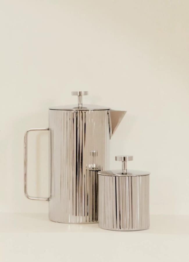 A tall, cylindrical metallic coffee pot with a handle stands beside a matching sugar bowl, both featuring vertical ridges and round lids, set against a plain light background—perfect examples of chic homeware under £50.