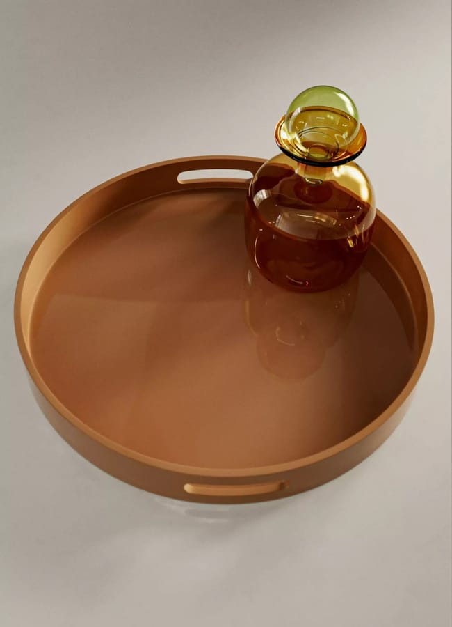 A round brown tray, reminiscent of elegant caramel homeware, holds a small, round glass bottle with a yellow-green stopper and sits on a light surface. The tray features two oval cut-out handles.