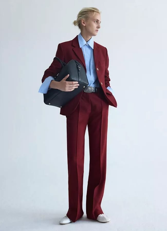 A person wearing a burgundy suit, light blue shirt, and white shoes holds a black bag under one arm, looking to the side against a plain light background—an embodiment of fashion essentials for an effortless wardrobe upgrade.