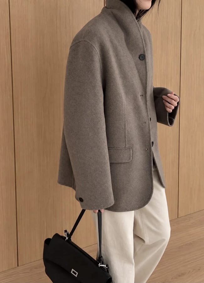 Person wearing a taupe oversized coat, ideal for layering over lightweight jackets, paired with cream trousers and holding a black handbag, standing in front of a wooden wall. Their face is partially out of frame.