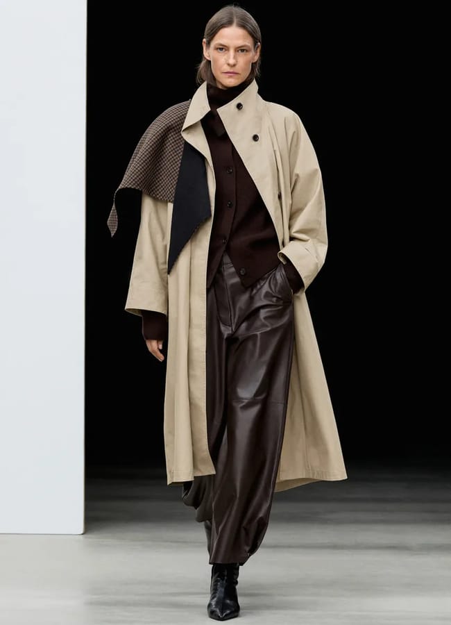 A model wears a beige trench coat—one of the ultimate wardrobe essentials—over a dark brown jacket and brown leather trousers, with a scarf draped over one shoulder, walking on a catwalk against a black background.