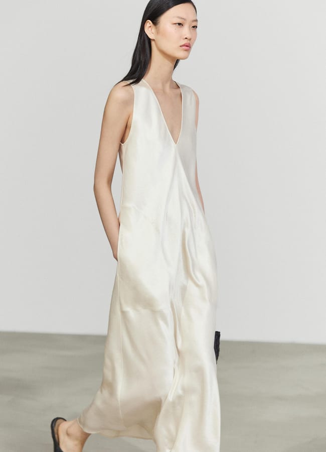 A person wearing a sleeveless, white, V-neck, ankle-length satin dress with side pockets—reminiscent of designs from minimalist high street brands—walks on a light grey floor against a plain white background.