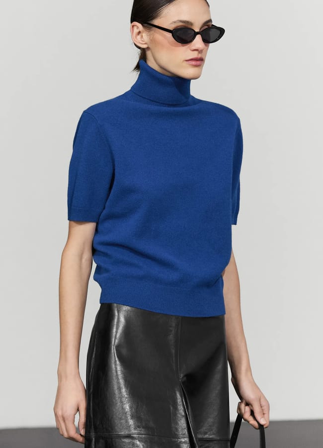 A woman wearing black sunglasses, a striking International Klein Blue short-sleeve polo-neck jumper, and a black leather skirt stands against a plain light grey background, bringing a bold pop of colour everywhere she goes.