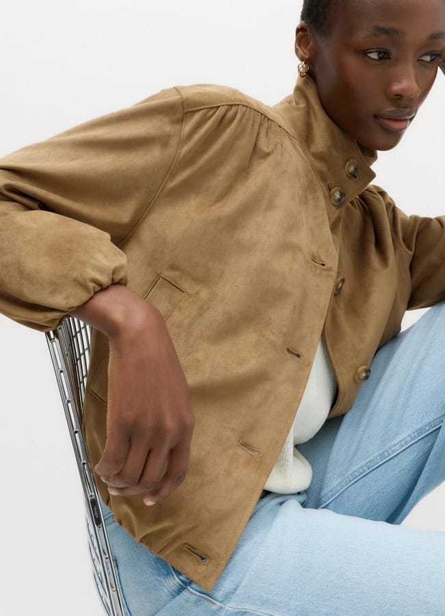A person wearing a tan lightweight jacket and light blue jeans sits on a metal chair, resting one arm on the chair’s back. The background is plain and light-coloured.