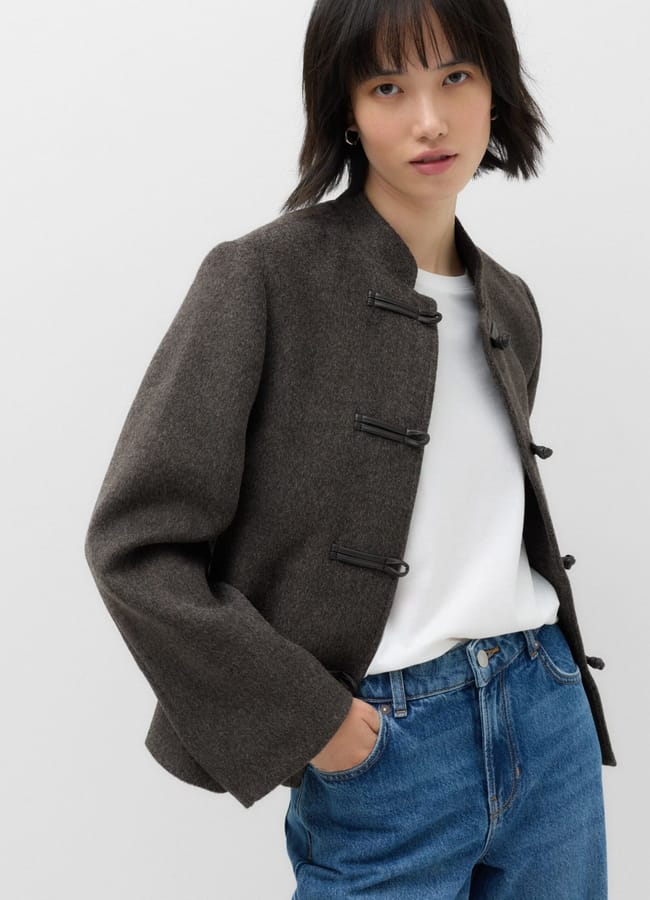 A person with short dark hair wears a grey mandarin-collar jacket over a white T-shirt and blue jeans, standing with one hand in their pocket against a plain light background, showcasing new season fashion.