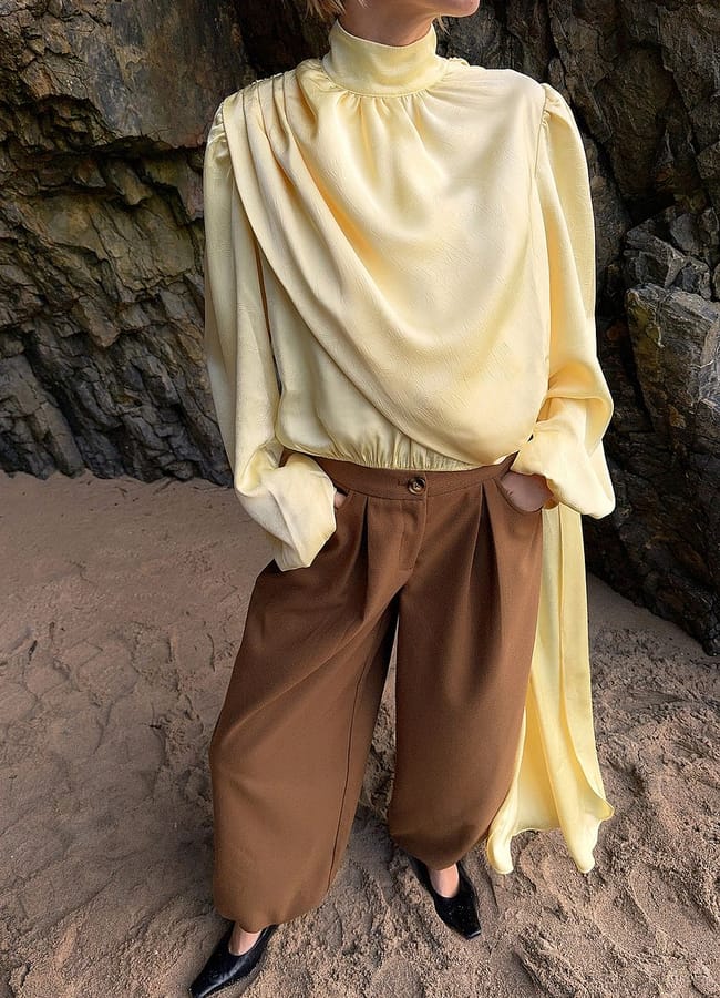 Person standing on sandy ground near rocks, wearing a loose yellow long-sleeve blouse with draped fabric and high neck, brown wide-leg trousers, and black shoes, with hands in pockets—a relaxed fashion statement that channels cool boyfriend clothes vibes.