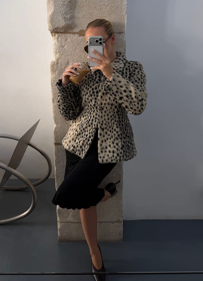 A woman in a spotted faux fur coat, one of this season’s must-have lightweight jackets, and a black skirt poses for a mirror selfie, holding an iced coffee. She stands on one leg, wearing black heels, with her hair tied back. Modern decor is visible in the background.